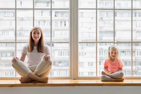 Junge Mutter beim Yoga mit 3-jähriger Tochter vor dem Fenster. glückliche Mutter lächelt, während sie zusammen mit ihrem süßen Mädchen Yoga praktiziert — Stockfoto