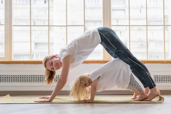 Ibu muda melakukan yoga dengan putri 3 tahun di depan jendela. Ibu bahagia tersenyum saat berlatih yoga bersama dengan gadis manisnya — Stok Foto
