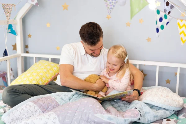 Closeup of young father dad in bed with his happy toddler daughter reading a book — ストック写真
