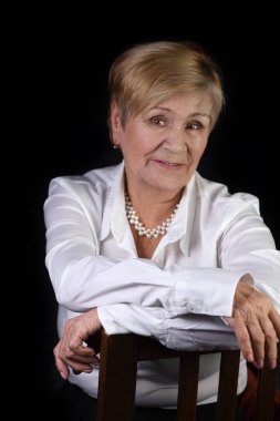 Bir sandalyede oturan yaşlı bir kadının (75 +) yarım boy portresi. Beyaz gömlek. Siyah arka plan. Yakın plan.