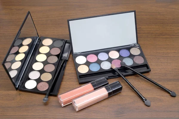 Opened cosmetic set with round colorful eye shadows and brushes in black plastic case with mirror and two pink lipstick tubes on wooden background closeup