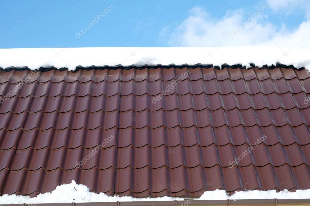 Side of country house roof from brown metal tile with snow in sunny spring day under blue sky with white clouds front view