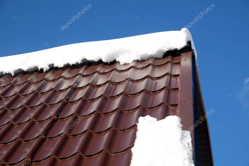 Side of country house roof from brown metal tile with snow in sunny spring day under blue sky with white clouds side view