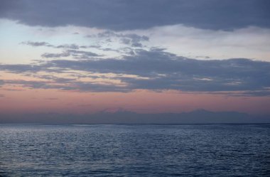 Yüksek dağ siluetlerinde alacakaranlık manzarası, ufukta beyaz kar örtüleri, gökyüzünün altında güzel bulutlarla denizden sonra ufuklarda.