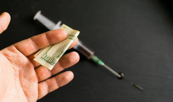 Money for drugs. A man\'s hand stretches dollars against the background of a syringe with drugs on a dark background.