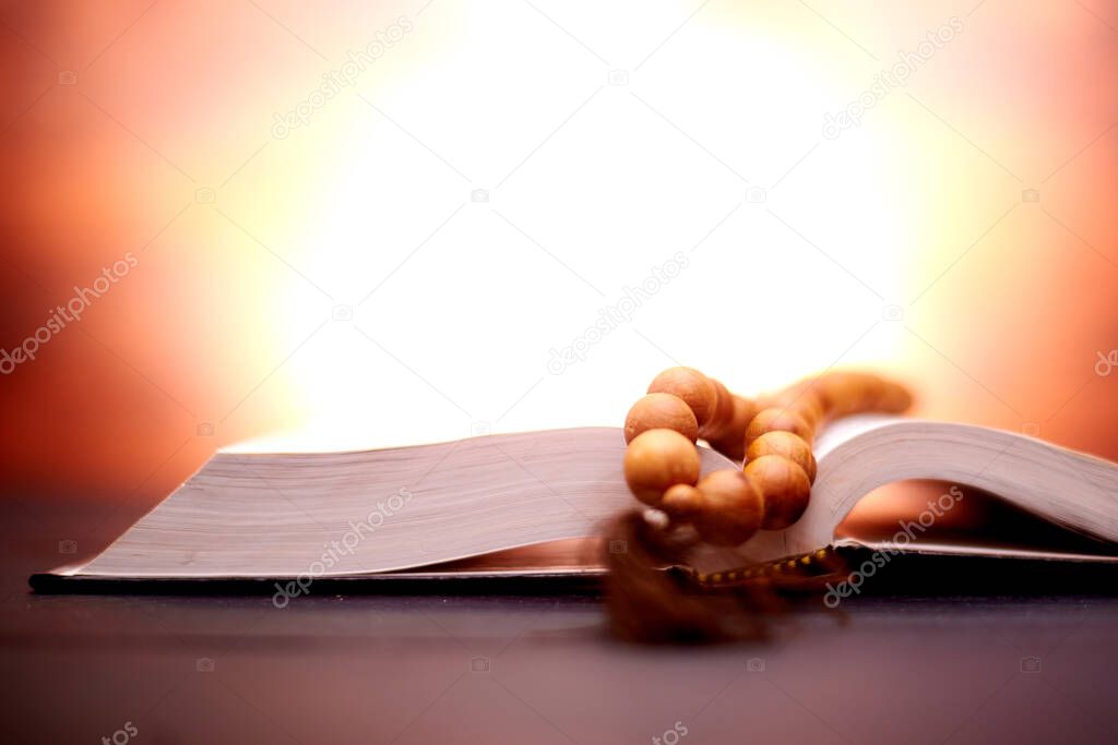 Bible and church rosary on a dark brown background with a radiance behind. Symbol of faith, religiosity, prayer.