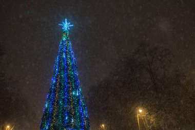 Noel ağacı süslenmiş sokak çelenkleri, gece parkta kar yağarken parlıyor..
