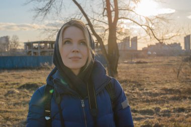 Gün batımında arka planda sıcak bir ceket giymiş bir kadın terk edilmiş binaların arka planına yaslanmış..