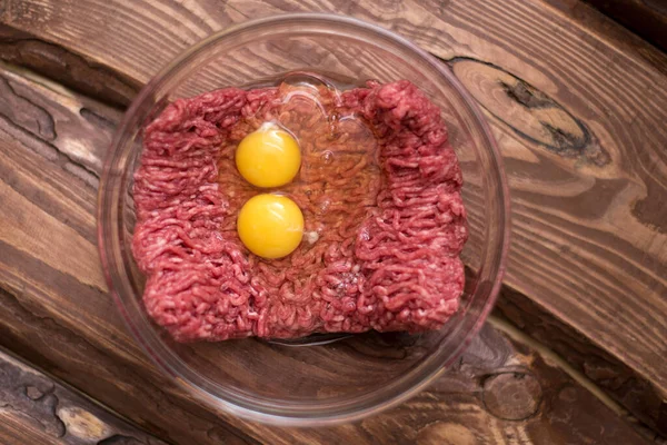 Fresh minced meat with chicken eggs on a plate on a wooden background for cooking.