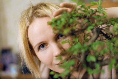 Bir kadın gülümsüyor ve bonsai ağacının yapraklarının arkasından dışarı bakıyor..