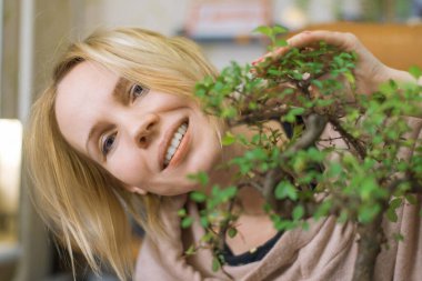 Bir kadın gülümsüyor ve bonsai ağacının yapraklarının arkasından dışarı bakıyor..