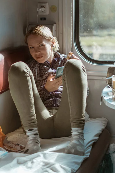 携帯電話を持っている女性はクッキーを食べ 電車のコンパートメントに座っています — ストック写真