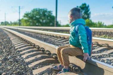 Sırt çantalı küçük bir çocuk raylarda oturuyor. Yürüyüş sırasında dinlenin. Treni bekliyorum..