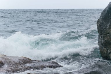 Dalgalar deniz kıyısındaki kayalara vuruyor, okyanus bulutlu havada..