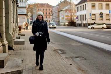 Paltolu bir kadın gezgin şehir caddesi boyunca elinde kamerayla yürüyor..