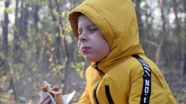 Bir çocuk piknikte ateşin yanında sosisli çörek yiyor. Duman ve küller yüzünde uçuşuyor..