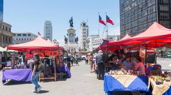 Mercado callejero con gente caminando, en la plaza Sotomayor, en el paseo marítimo de Valparaíso, en Chile. Al fondo, monumento al héroe Arturo Prat . — Foto de Stock