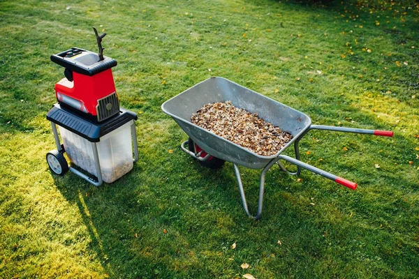 Astilladora de jardín, trituradora eléctrica (mulcher) con carretilla llena de mantillo de madera, fondo de hierba verde — Foto de Stock