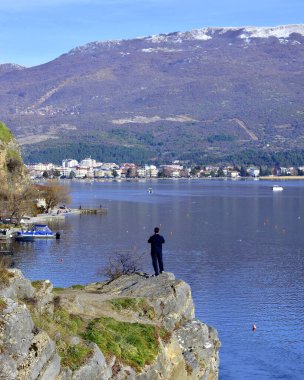 OHRID, MACEDONIA - FEBRUARY 23, 2020: Man on the cliff picturing Lake Ohrid and city of Ohrid with smartphone