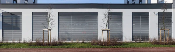 Near the long wall of the city garage with latticed  windows, young rowan trees are planted. Sunny day panoramic natural colors simplistic panoramic collage