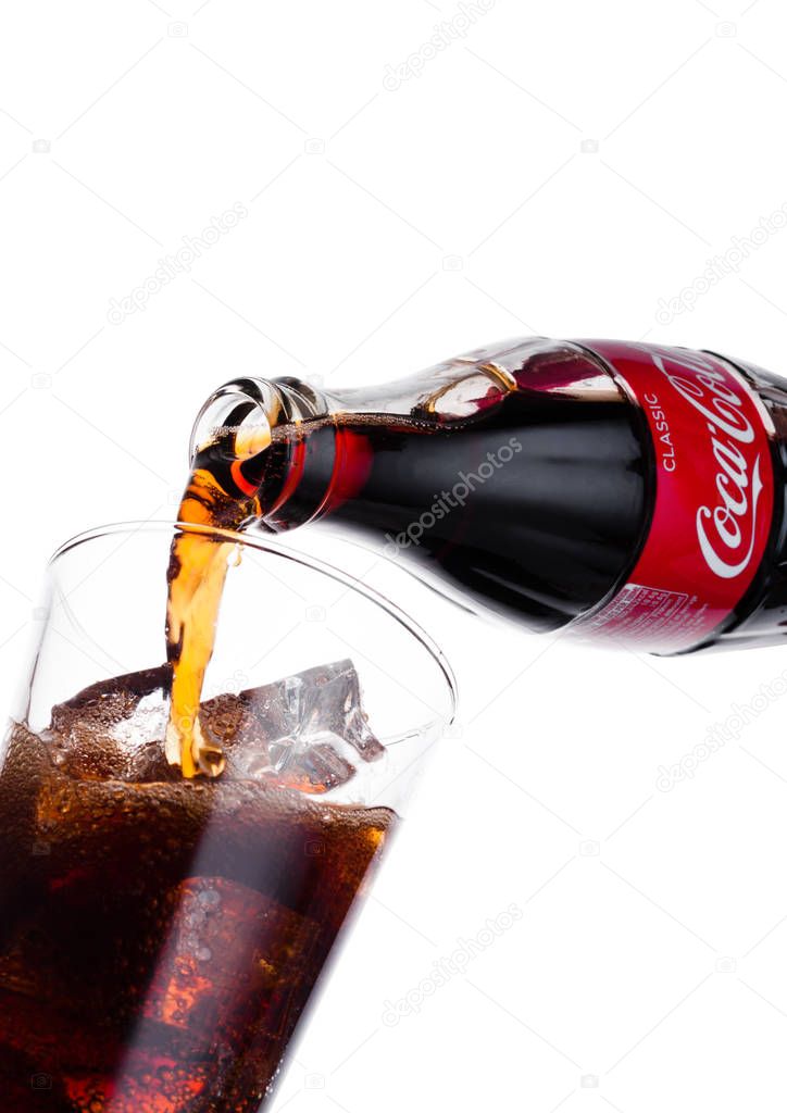 LONDON, UK - JANUARY 02, 2018: Pouring Coca Cola soda drink from bottle to glass on white background. The drink is produced and manufactured by The Coca-Cola Company.