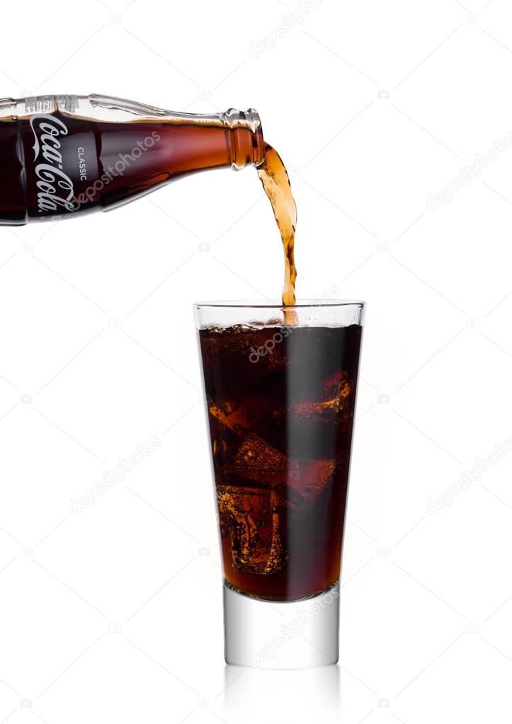 LONDON, UK - JANUARY 02, 2018: Pouring Coca Cola soda drink from bottle to glass on white background. The drink is produced and manufactured by The Coca-Cola Company.