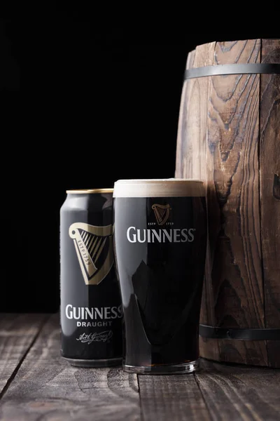 London, Verenigd Koninkrijk - 03 mei 2018: Aluminium blikje en originele glas Guinness bier van het vat stout naast houten vat op donkere houten achtergrond. — Stockfoto