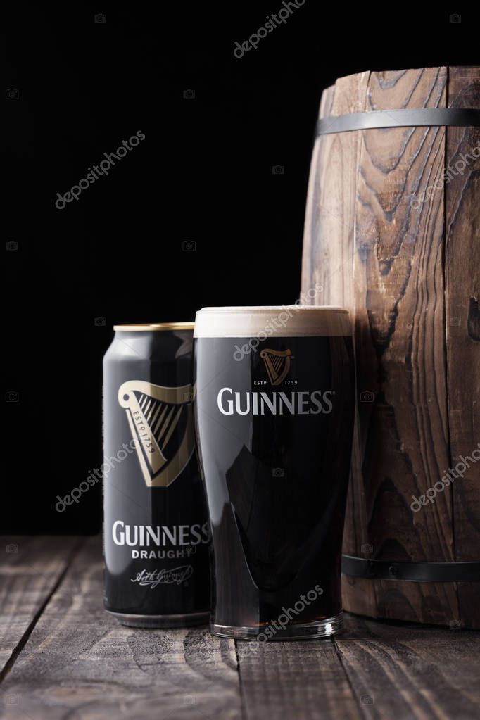 LONDON, UK - MAY 03, 2018: Aluminium can and original glass of Guinness draught stout beer next to wooden barrel on dark wooden background. Guinness beer has been produced since 1759 in Dublin.