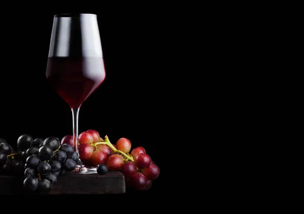 Copo elegante de vinho tinto com uvas escuras e vermelhas em tábua de madeira sobre fundo preto. Espaço para texto — Fotografia de Stock