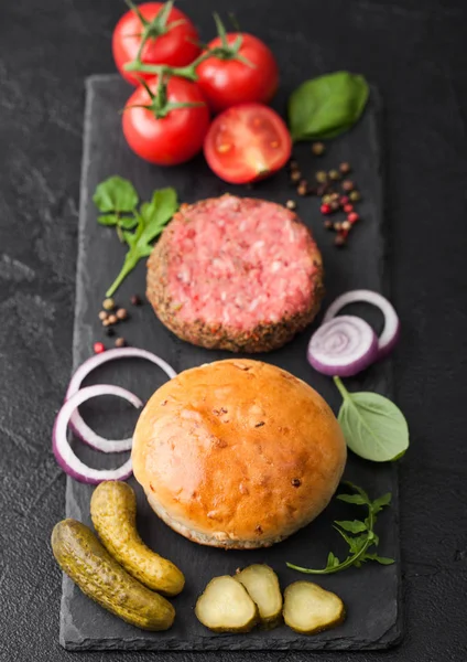 Hamburguesa fresca de carne picada de pimienta cruda sobre tabla picadora de piedra con bollos de cebolla y tomates sobre fondo negro. Pepinillos salados y albahaca . — Foto de Stock