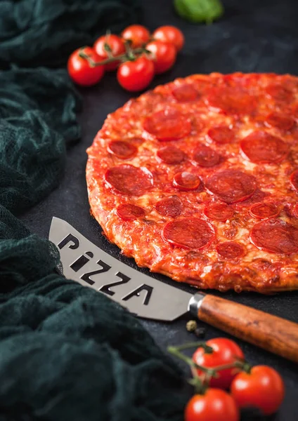 Fresh round baked Hot and Spicy Pepperoni pizza with knife and tomatoes with basil on black background with kitchen cloth. Close up