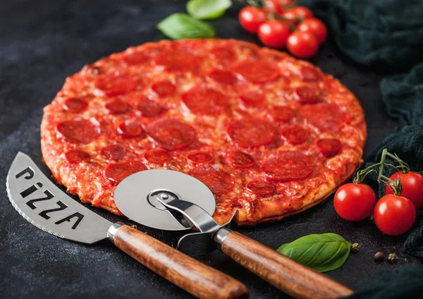 Fresh round baked Hot and Spicy Pepperoni pizza with wheel cutter and knife with tomatoes and basil on black background with kitchen cloth. Close up