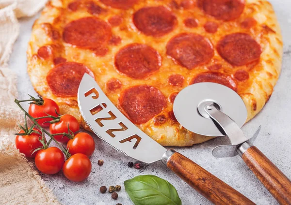 Fresh round baked Pepperoni italian pizza with wheel cutter and knife with tomatoes and basil on light background with linen towel. Macro