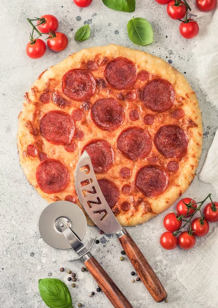 Fresh round baked Pepperoni italian pizza with wheel cutter and knife with tomatoes and basil on light background with linen towel. Top view