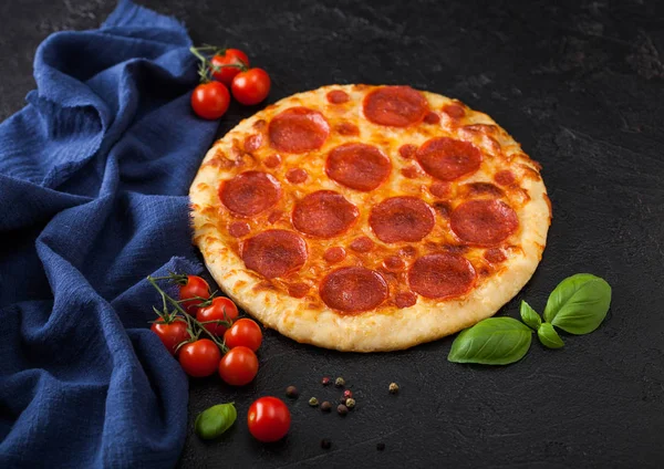Fresh round baked Pepperoni italian pizza with tomatoes with basil on black background with blue linen towel.