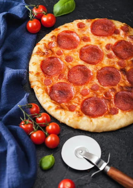 Fresh round baked Pepperoni italian pizza with wheel cutter and tomatoes with basil on black background. Top view