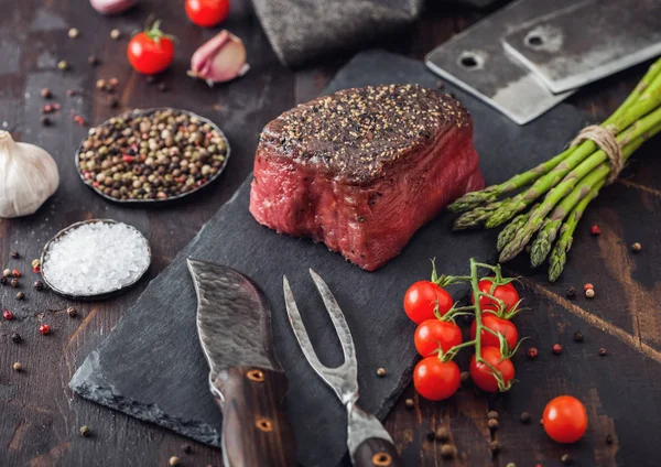 Slice of Raw Beef Topside Joint with Salt and Pepper on stone chopping board with fork and knife, garlic and asparagus tips and tomatoes. — Stock Photo, Image