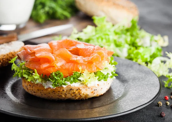 Sandwich de bagel fresco y saludable con salmón, ricotta y lechuga en plato negro sobre fondo de mesa de cocina negro. Alimento saludable para adelgazar. Vaso de leche y verduras frescas — Foto de Stock