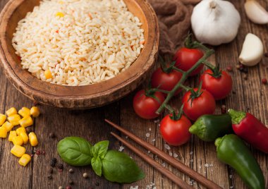 Ahşap kase, haşlanmış uzun tahıllı basmati pilavı, ahşap masa arkasında sebzeler, çubuklar, domatesler, mısır, sarımsak ve fesleğen. Makro