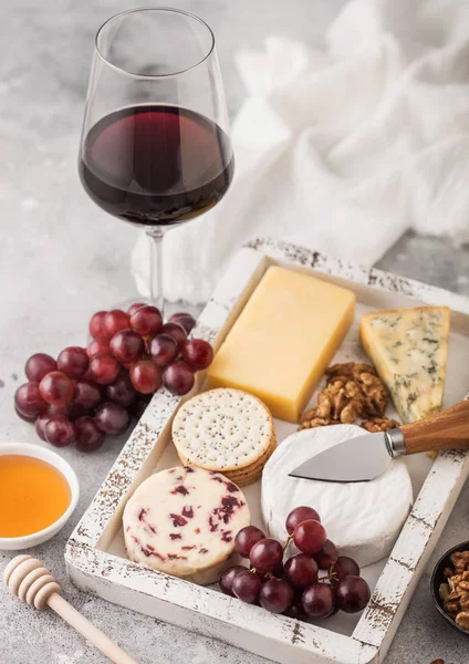 Rotweinglas mit verschiedenen Käsesorten in Holzkiste und Trauben auf hellem Tischhintergrund. Blue Stelton, Red Leicester und Brie-Käse mit Cheddar und Nüssen mit Honig. — Stockfoto