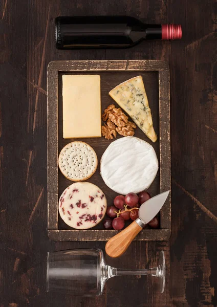 Glas und Flasche Rotwein mit verschiedenen Käsesorten in Holzkiste und Trauben auf Holztischhintergrund. blue stilton, red leicester und brie cheese. Ansicht von oben — Stockfoto