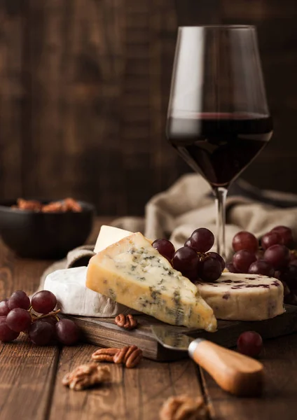 Tahtasında çeşitli peynirler ve ahşap zemin üzerinde üzümler olan bir bardak kırmızı şarap. Mavi Stilton, Kırmızı Leicester ve Brie Cheese ve bir kase fındık.. — Stok fotoğraf