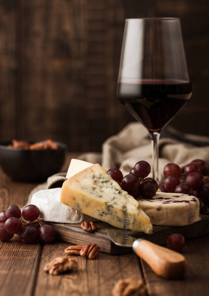Glass of red wine with selection of various cheese on the board and grapes on wooden background. Blue Stilton, Red Leicester and Brie Cheese and bowl of nuts.