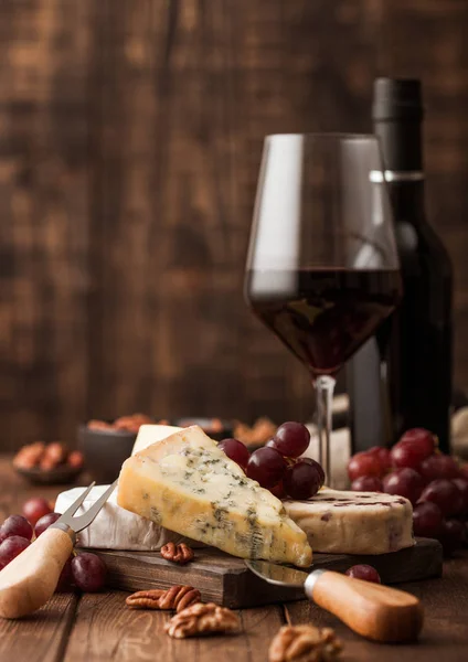 Bicchiere e bottiglia di vino rosso con selezione di formaggi vari sul bordo e uva su fondo di legno. Blue Stilton, Red Leicester e Brie Cheese e ciotola di noci . — Foto Stock