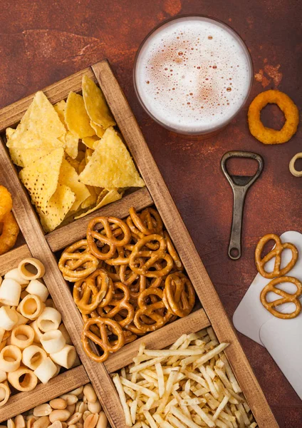 Glass of craft lager beer and opener with box of snacks on brown background. Pretzel,salty potato sticks, peanuts, onion rings with nachos in vintage box with openers and beer mats. Top view — Stock Photo, Image