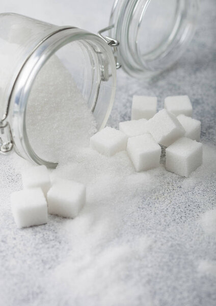 Glass jar of natural white refined sugar with cubes on light table background.