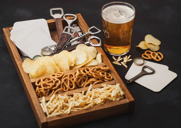 Bicchiere di birra artigianale lager e apri con scatola di snack su sfondo nero. Pretzel e patatine e bastoncini di patate salate in scatola di legno vintage con apriscatole e stuoie di birra. Macro — Foto Stock