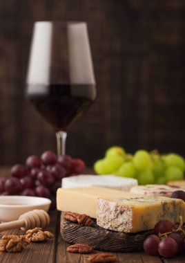 Tahtasında çeşitli peynirler ve ahşap zemin üzerinde üzümler olan bir bardak kırmızı şarap. Mavi Stilton, Kırmızı Leicester ve Brie Cheese ve bir kase fındık ve bal.