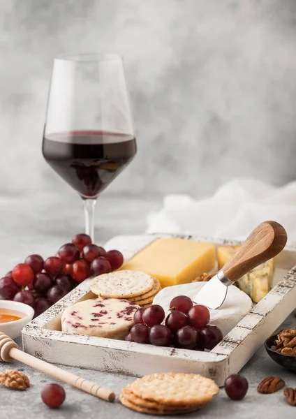 Rotweinglas mit verschiedenen Käsesorten in Holzkiste und Trauben auf hellem Tischhintergrund. Blue Stelton, Red Leicester und Brie-Käse mit Cheddar und Nüssen mit Honig. — Stockfoto