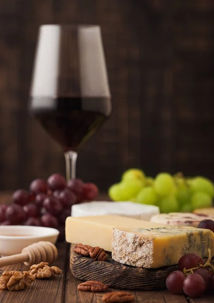 Tahtasında çeşitli peynirler ve ahşap zemin üzerinde üzümler olan bir bardak kırmızı şarap. Mavi Stilton, Kırmızı Leicester ve Brie Cheese ve bir kase fındık ve bal. — Stok fotoğraf